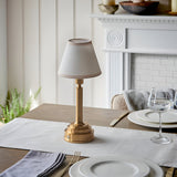 Danebourne LED rechargeable table lamp in brass with cream fabric shade, styled on a dining table beside white plates and a glass, near a white fireplace.