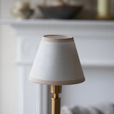 Danebourne LED Rechargeable Table Lamp with tapered fabric shade in white and brass, on a stepped brass base, set against a blurred neutral interior.