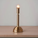 Danebourne LED rechargeable table lamp in brass with slim column, stepped base and touch dimmer, glowing on a wooden surface against a grey background.