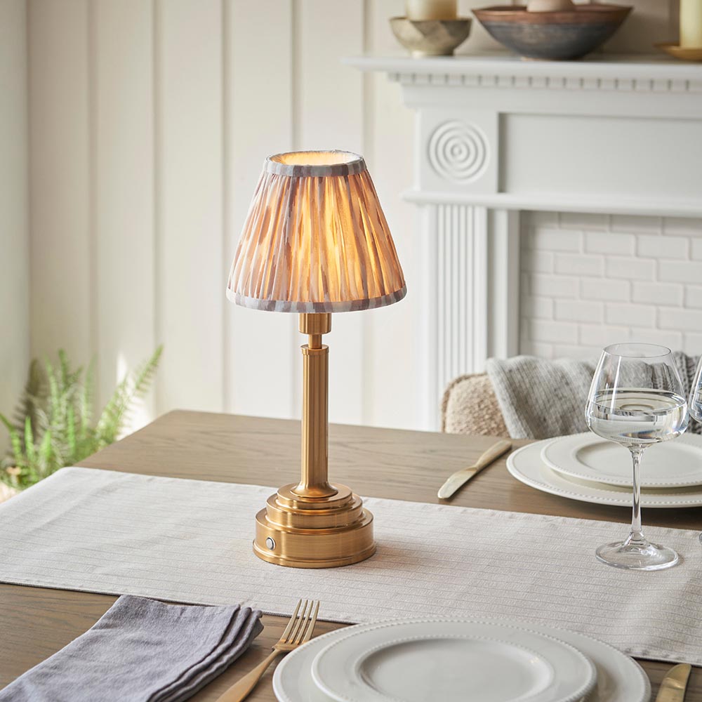 Danebourne LED Ikat Table Lamp in brass with a pleated ikat-print shade, shown lit on a dining table beside white plates and glassware near a fireplace.