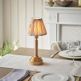 Danebourne LED Ikat Table Lamp in brass with a pleated ikat-print shade, shown lit on a dining table beside white plates and glassware near a fireplace.