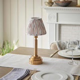 Danebourne LED Ikat table lamp with brass base and pleated pink and white Ikat fabric shade, shown on a dining table beside white plates and a wine glass.