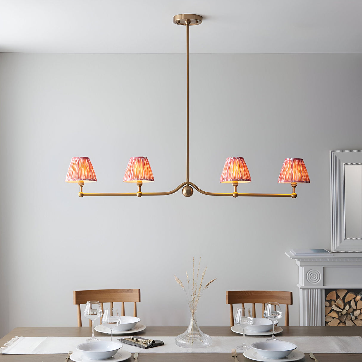 Endon Cliffleigh 4 light linear pendant in brushed aged brass with four coral pink pleated ikat taper shades, shown above a dining table.