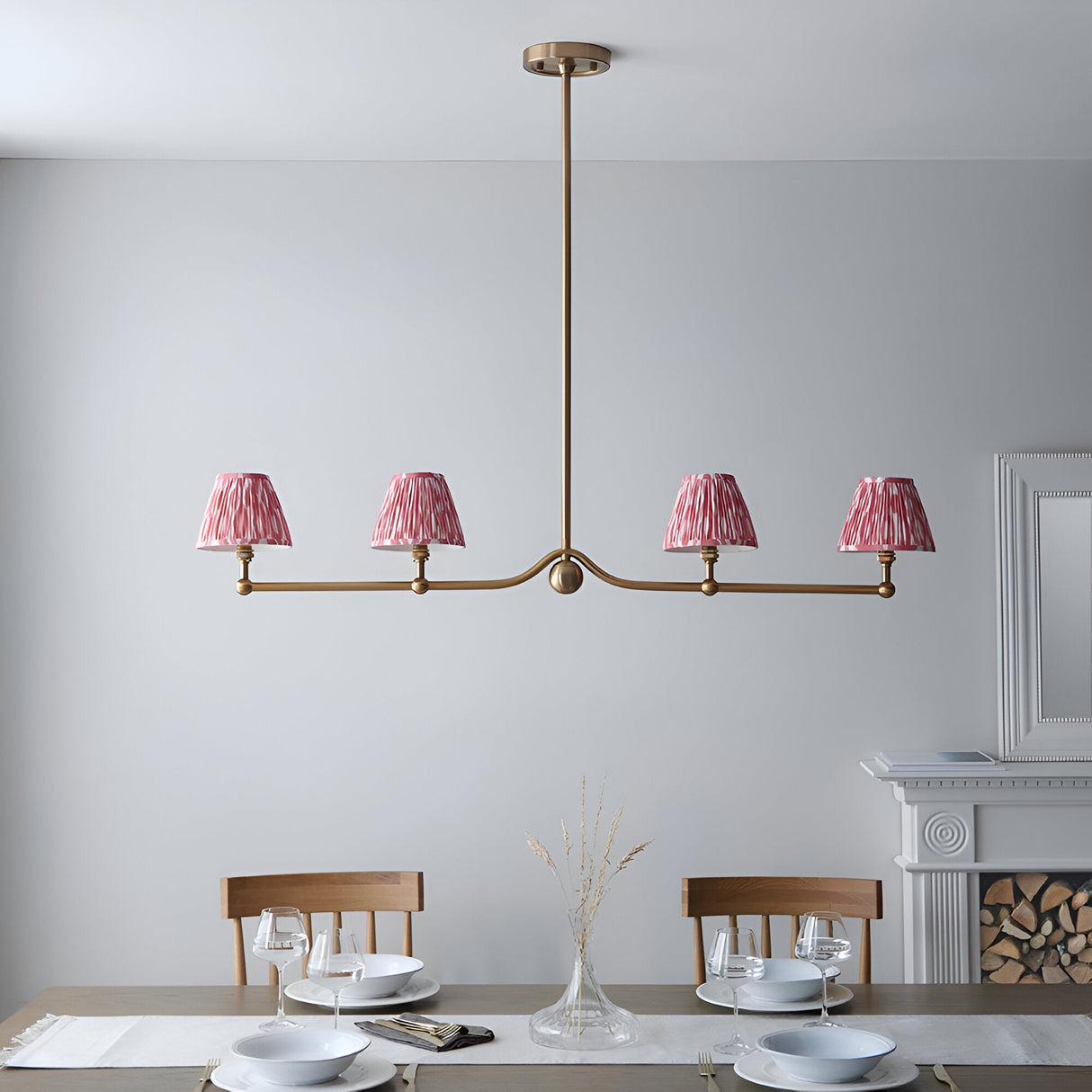 Endon Cliffleigh 4 light linear pendant in brushed aged brass with coral pink pleated ikat tapered shades, hung above a dining table by a fireplace.