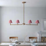Endon Cliffleigh 4 light linear pendant in brushed aged brass with coral pink pleated ikat tapered shades, hung above a dining table by a fireplace.