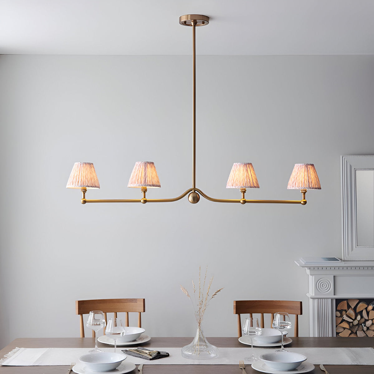 Cliffleigh Leaf 4 Light Linear Pendant in brushed brass with four pleated fabric shades on a slim bar, shown above a dining table in a light-toned room.