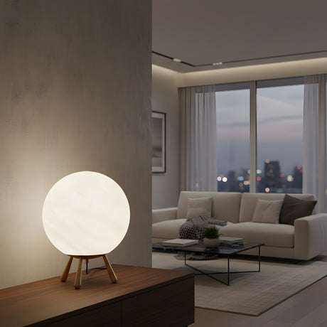 The Orlena Small Globe Alabaster Table Lamp in Brass & Soft White sits on a wooden sideboard in a modern living room with a white sofa, glass coffee table, and large windows overlooking the city at dusk.