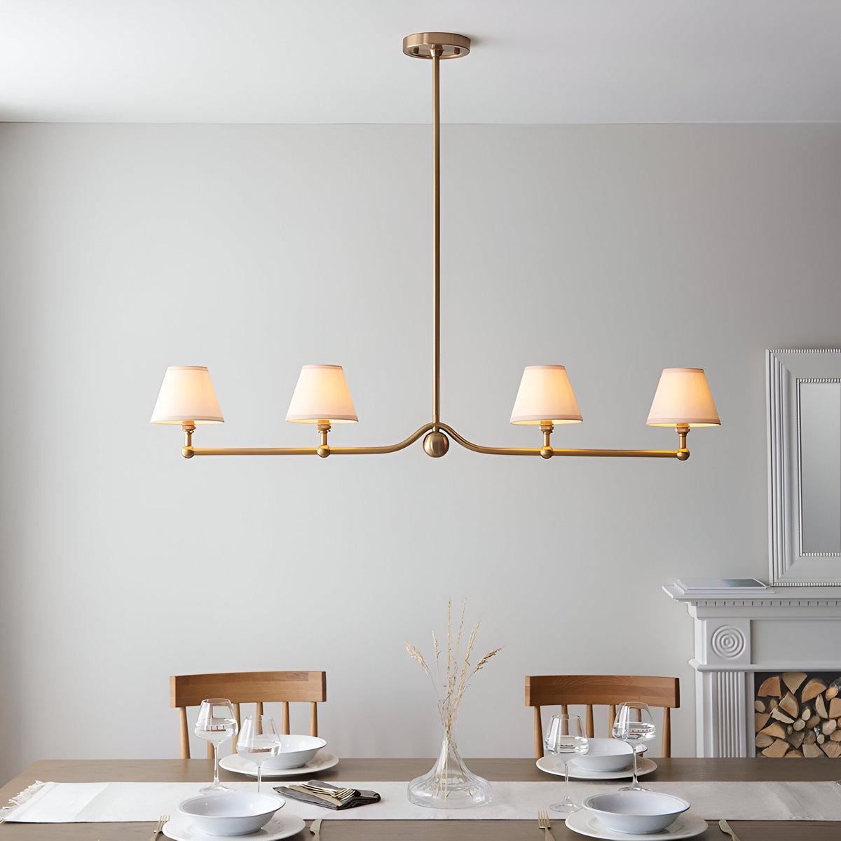 Endon Cliffleigh 4 light linear pendant in brushed aged brass with white tapered fabric shades, shown above a dining table near a fireplace.