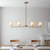 Endon Cliffleigh 4 light linear pendant in brushed aged brass with white tapered fabric shades, shown above a dining table near a fireplace.