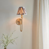 Endon Cliffleigh Zigzag brushed aged brass wall light with a tapered pleated pearl grey zigzag shade, mounted beside a cream curtain above foliage.