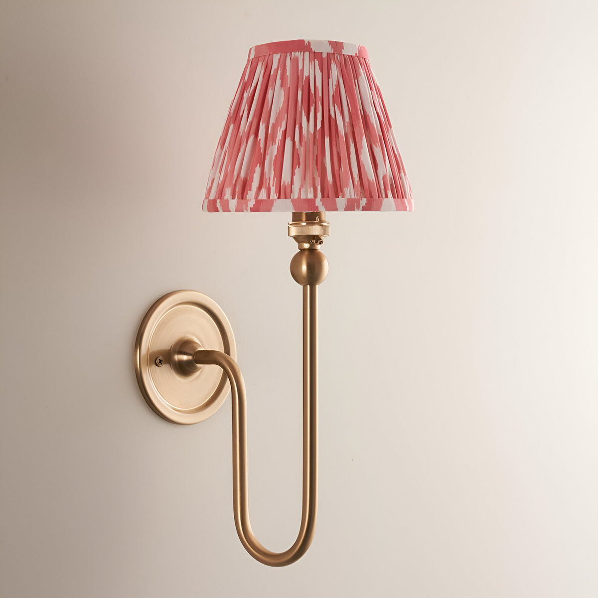 Endon Cliffleigh Ikat wall light in brushed brass with a tall curved arm and pleated pink Ikat-print tapered shade, on a plain pale background.