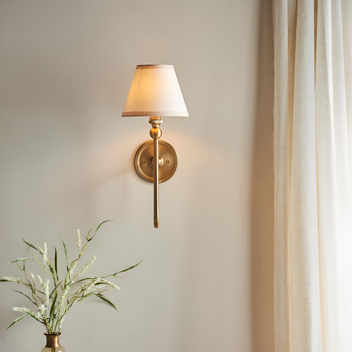 Cliffleigh Ivy wall light in brushed aged brass with a tapered vintage white fabric shade, shown illuminated on a bedroom wall beside a curtain.