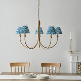 Endon Cliffleigh 5 light pendant with curved brushed brass arms and tapered marlin blue ripple fabric shades, hung over a dining table by a fireplace.