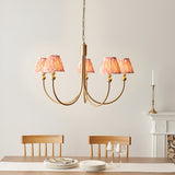 Cliffleigh 5-Light Curved Arm Ikat Pendant in Coral with brushed aged brass frame and pleated fabric shades, above a dining table with wooden chairs.
