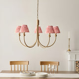 Cliffleigh 5 Light Curved Arm Ikat Pendant in brushed aged brass with coral pink pleated fabric shades, shown above a dining table.