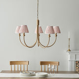 Cliffleigh Leaf 5 light curved arm pendant in brushed brass with pleated pink patterned shades, hung above a dining table in a light-toned room.