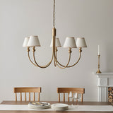 Cliffleigh 5 Light Curved Arm Pendant with brushed aged brass frame and tapered white fabric shades, shown above a dining table in a neutral-toned room.