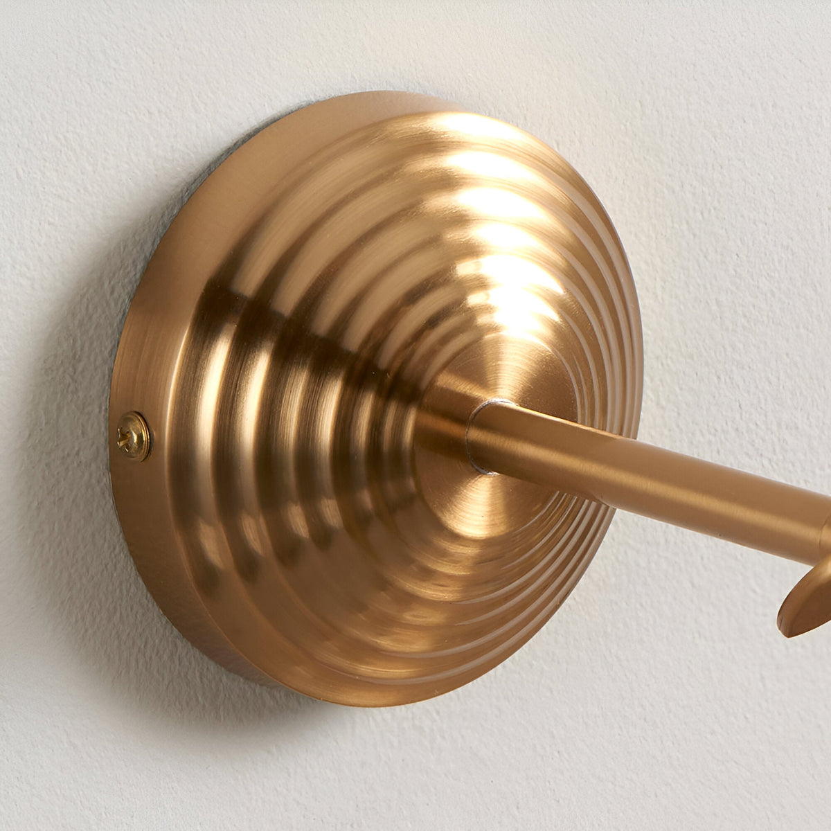 Close-up of the Endon Castley wall light's stepped circular back plate and linear arm in brushed aged brass, mounted on a white wall.