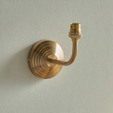Castgrove Wall Light in brushed aged brass with a stepped circular backplate, curved arm and B22 lampholder, shown against a plain light grey wall.