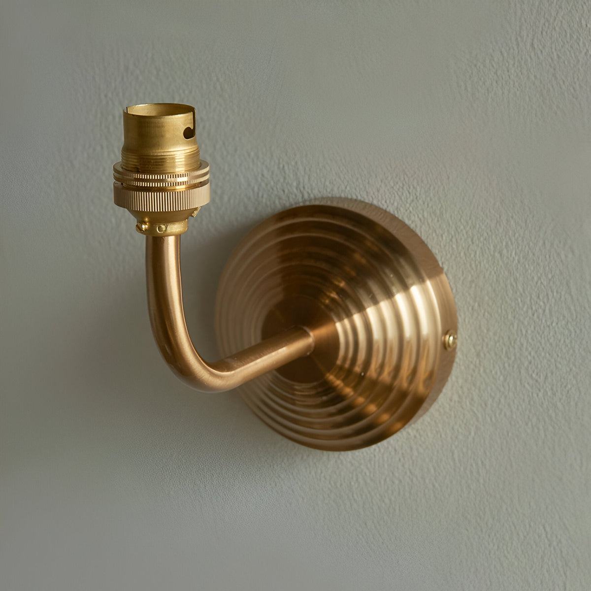 Castgrove wall light in brushed aged brass, with a curved arm and stepped circular backplate holding a B22 lampholder, shown against a plain grey wall.