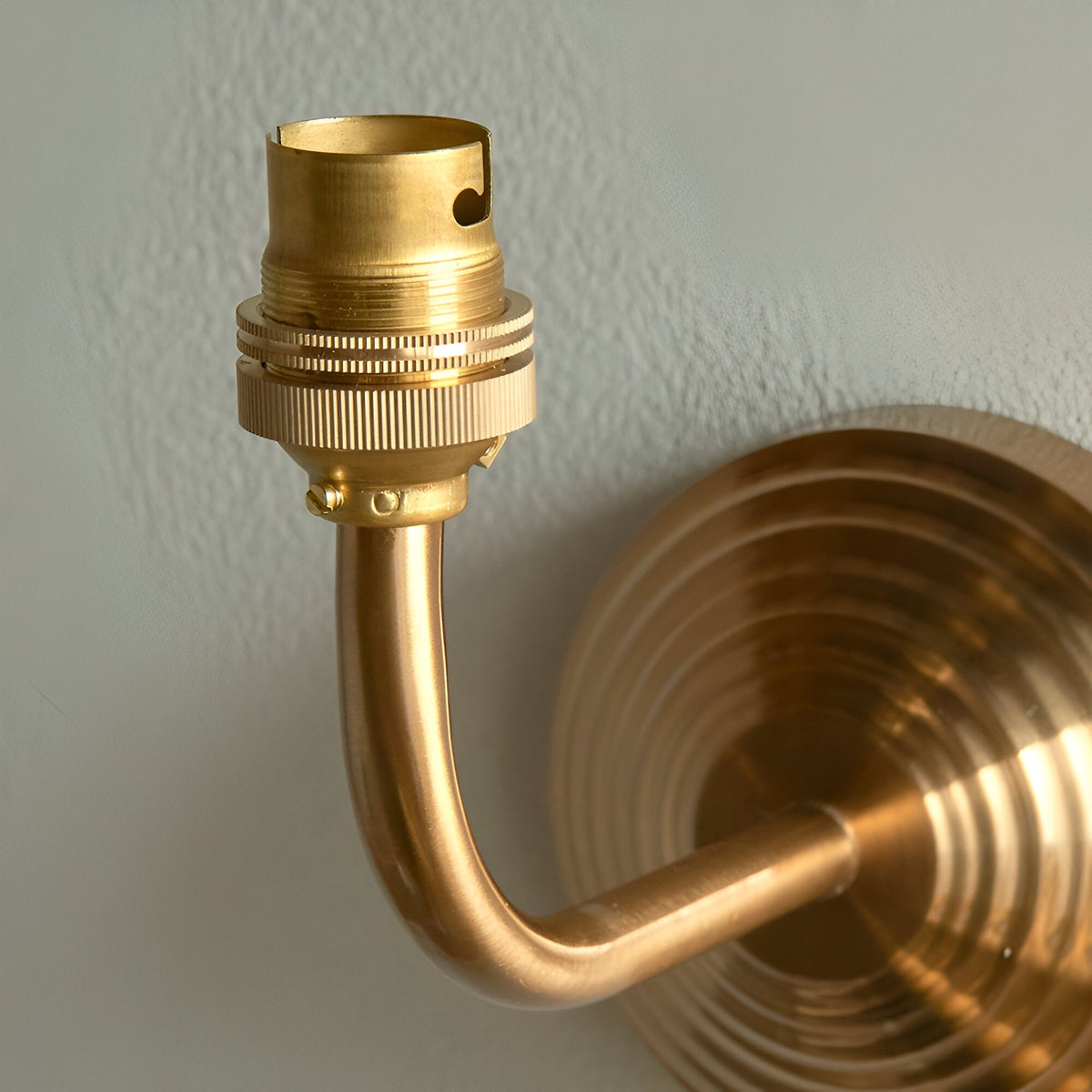 Castgrove wall light in brushed aged brass, close-up of the curved arm and B22 lampholder against a stepped circular wall backplate.