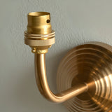 Castgrove wall light in brushed aged brass, close-up of the curved arm and B22 lampholder against a stepped circular wall backplate.
