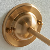 Close-up of Endon Brackdale wall light backplate in brushed aged brass, showing the domed mount, curved arm and knurled fixing screws on white wall.