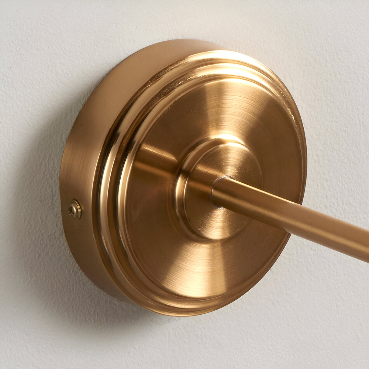 Close-up of the Endon Corven wall light's stepped circular backplate and linear arm in brushed aged brass, mounted on a plain white wall.