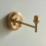 Endon Cliffwood wall light in brushed aged brass, with a stepped round backplate, adjustable knuckle, linear arm, and B22 lampholder on a pale wall.