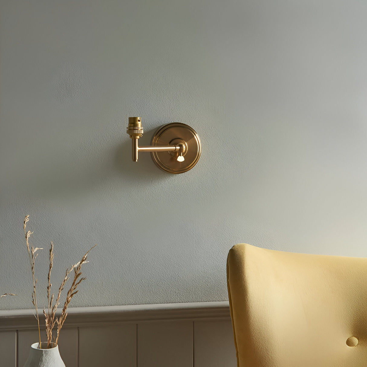 Endon Cliffwood brushed aged brass wall light with stepped back plate, adjustable knuckle arm and B22 lampholder, mounted on a grey wall by a yellow armchair.