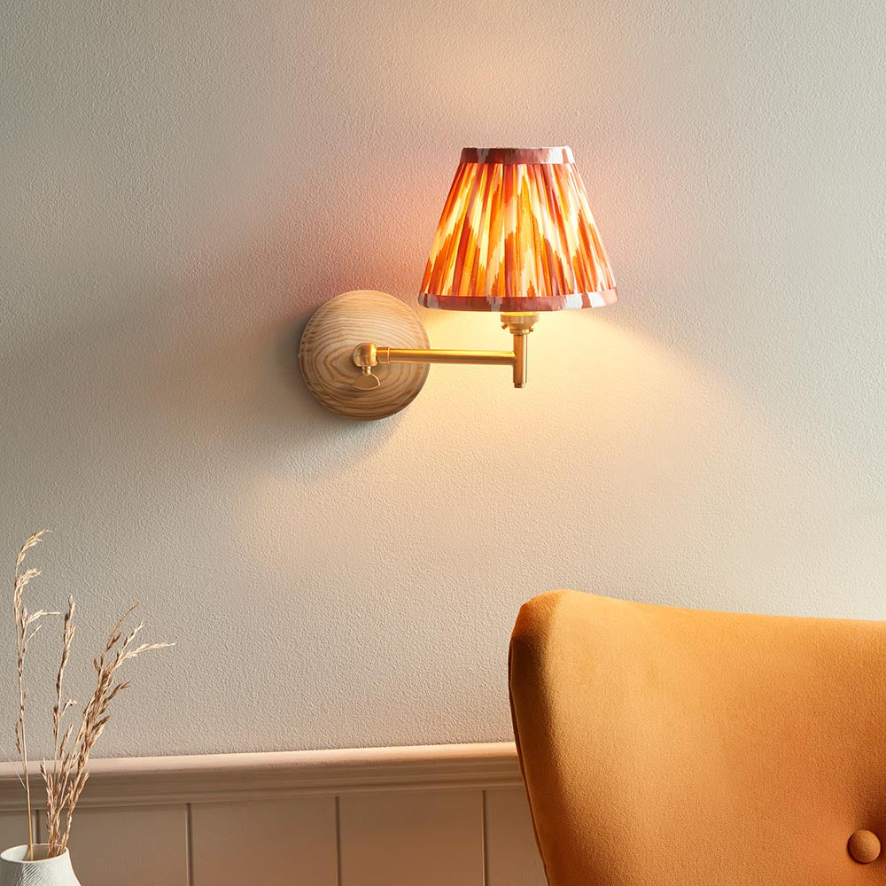 Endon Daneley wall light with natural ash wood back and brushed aged brass folding arm, topped with a pink and orange pleated shade above a mustard chair.