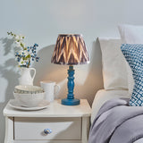 Aldworth Zigzag Table Lamp with turned blue wooden base and patterned zigzag fabric shade, shown lit on a bedside table beside crockery and bedding.