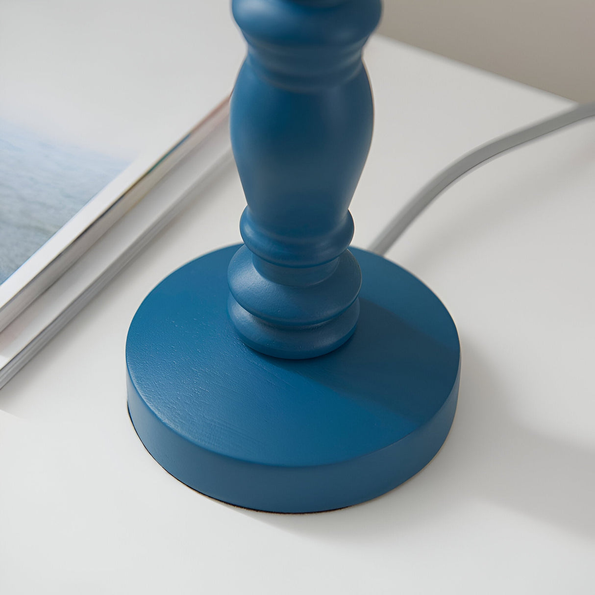 Close-up of the Aldworth Table Lamp's wooden base in blue, showing the turned spindle stem and round matte finish on a white surface.