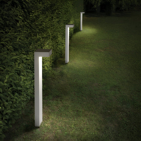 A row of Luma Outdoor Bollard Lights - White, standing 80cm tall, softly illuminates a grassy pathway at night, complemented by a lush green hedge on the left side of the image.