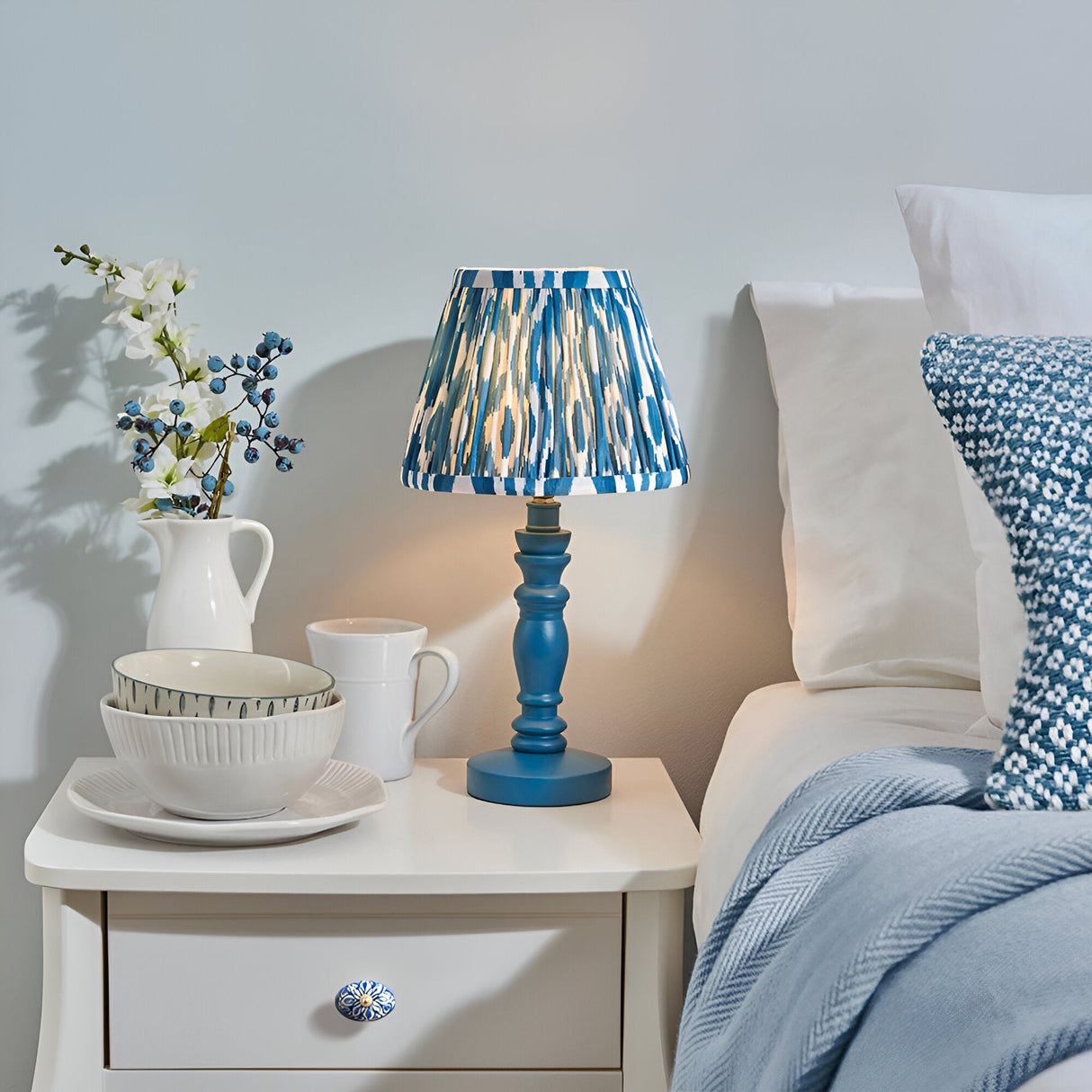 Endon Aldworth Ikat table lamp with marlin blue turned wood spindle base and pleated blue ikat shade, on a white bedside table beside a bed.