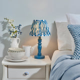 Endon Aldworth Ikat table lamp with marlin blue turned wood spindle base and pleated blue ikat shade, on a white bedside table beside a bed.
