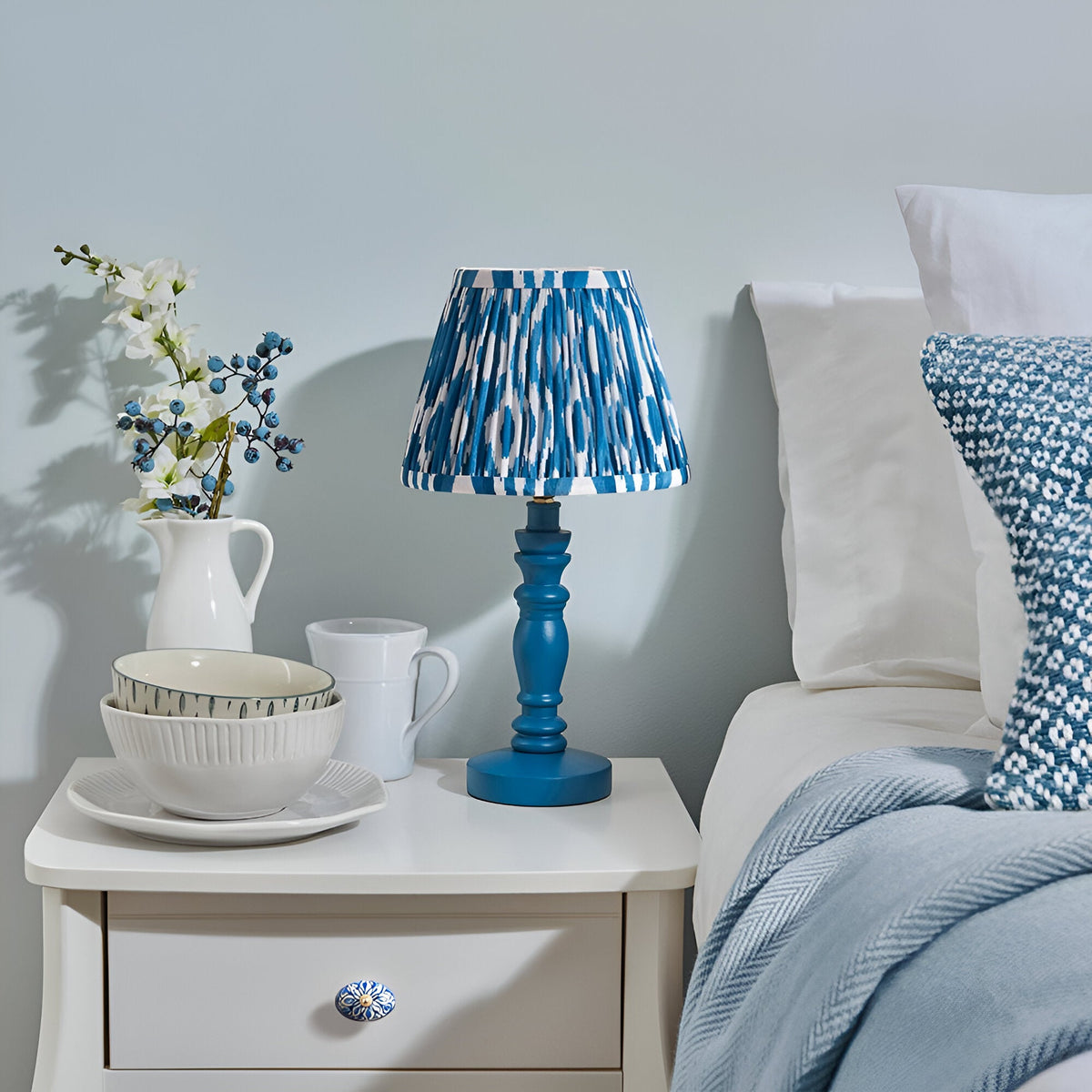 Endon Aldworth Ikat table lamp with marlin blue turned wooden spindle base and pleated blue ikat shade on a white bedside drawer beside a bed.