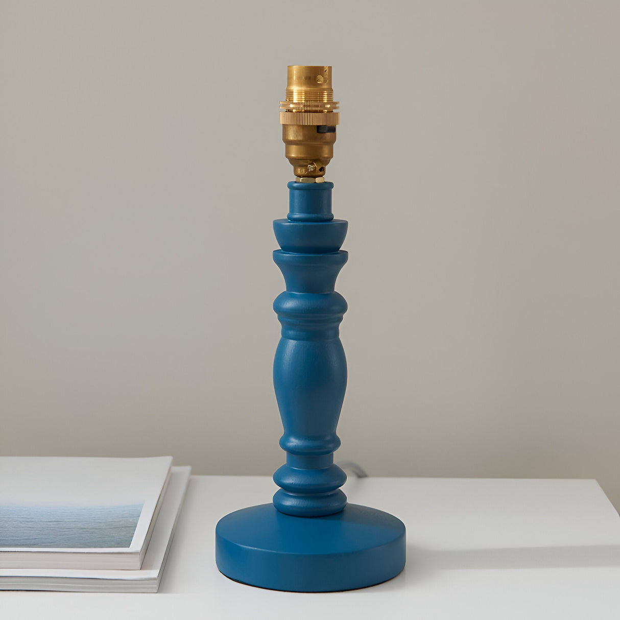 Endon Aldworth turned wooden spindle table lamp base in marlin blue with a brass B22 switched lampholder, shown on a white surface beside a book.