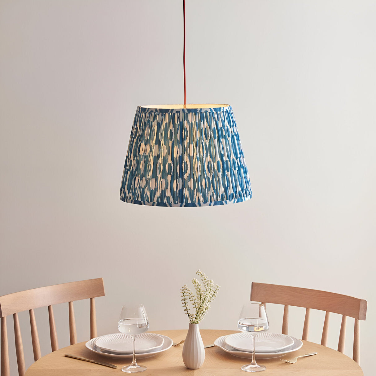 Endon Brackmont Ikat pendant light with pleated blue faux silk tapered shade and red fabric cable, hung above a wooden dining table set for two.