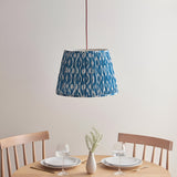 Endon Brackmont Ikat pendant with pleated marlin blue faux silk tapered shade on a red fabric cable, hung above a wooden dining table set for two.