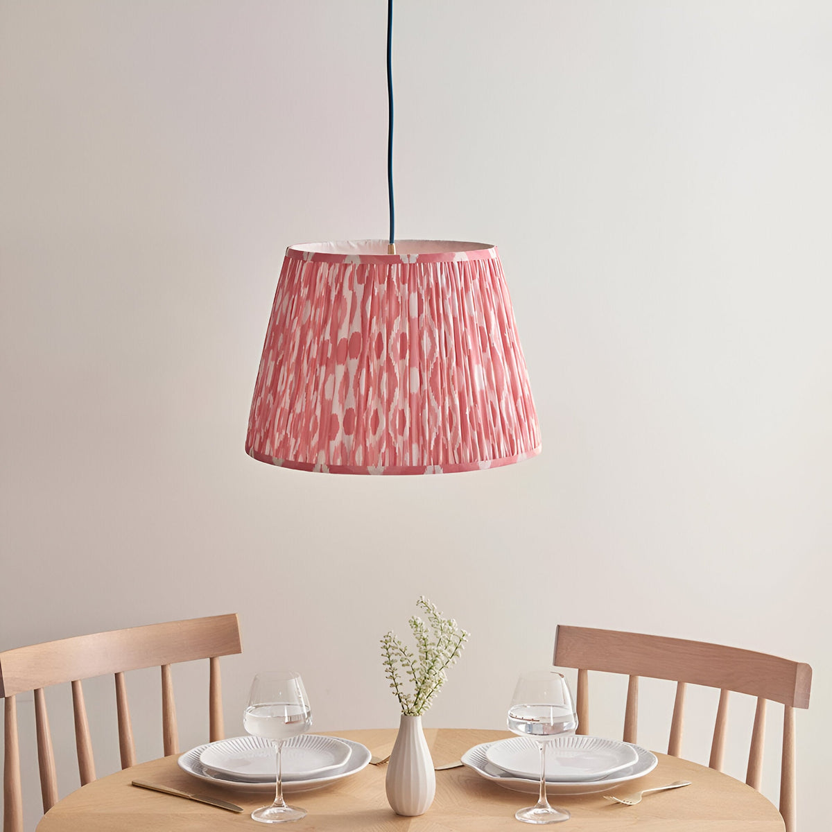 Endon Brackmont Ikat pendant with coral pink pleated faux silk tapered shade on a blue fabric cable, hanging above a wooden dining table set for two.