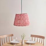 Endon Brackmont Ikat pendant with coral pink pleated faux silk tapered shade on a blue fabric cable, hanging above a wooden dining table set for two.