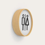 Bloom Signage Symbol lighting accessory in matt black iron, round wall light with wood-toned rim showing a cut-out male and female toilet symbol, white background.