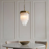 Aldfield Pendant in brushed aged brass, tiered clear glass rods over a single light, hung above a dining table against panelled grey walls.