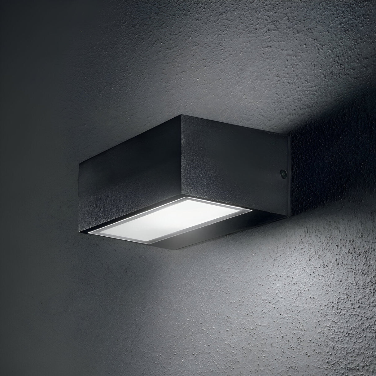 The Prism Outdoor Wall Light - Black, a sleek and contemporary wall-mounted fixture crafted from die-cast aluminum, illuminates the gray textured wall. It emits a soft glow that highlights the surface of the wall.