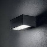 The Prism Outdoor Wall Light - Black, a sleek and contemporary wall-mounted fixture crafted from die-cast aluminum, illuminates the gray textured wall. It emits a soft glow that highlights the surface of the wall.