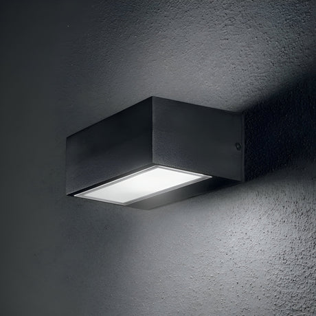 The Prism Outdoor Wall Light - Black, a sleek and contemporary wall-mounted fixture crafted from die-cast aluminum, illuminates the gray textured wall. It emits a soft glow that highlights the surface of the wall.