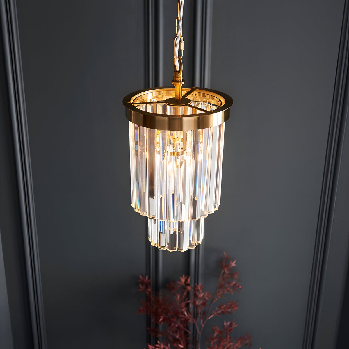 Endon Bevstead small single pendant with tiered clear lozenge glass drops on a brushed aged brass frame, hung against a dark panelled wall.