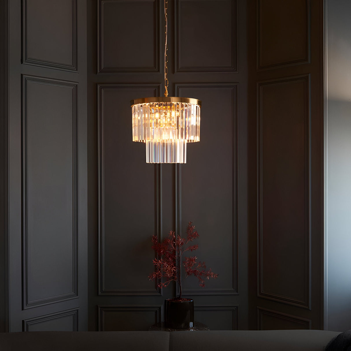 Bevstead 4-light pendant in brass with tiered clear glass drops, hanging lit in a dark panelled room above a small side table.
