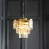 Endon Bevstead 7 light pendant in brushed brass with tiered clear cut glass drops, hung from a chain against dark grey panelled walls.